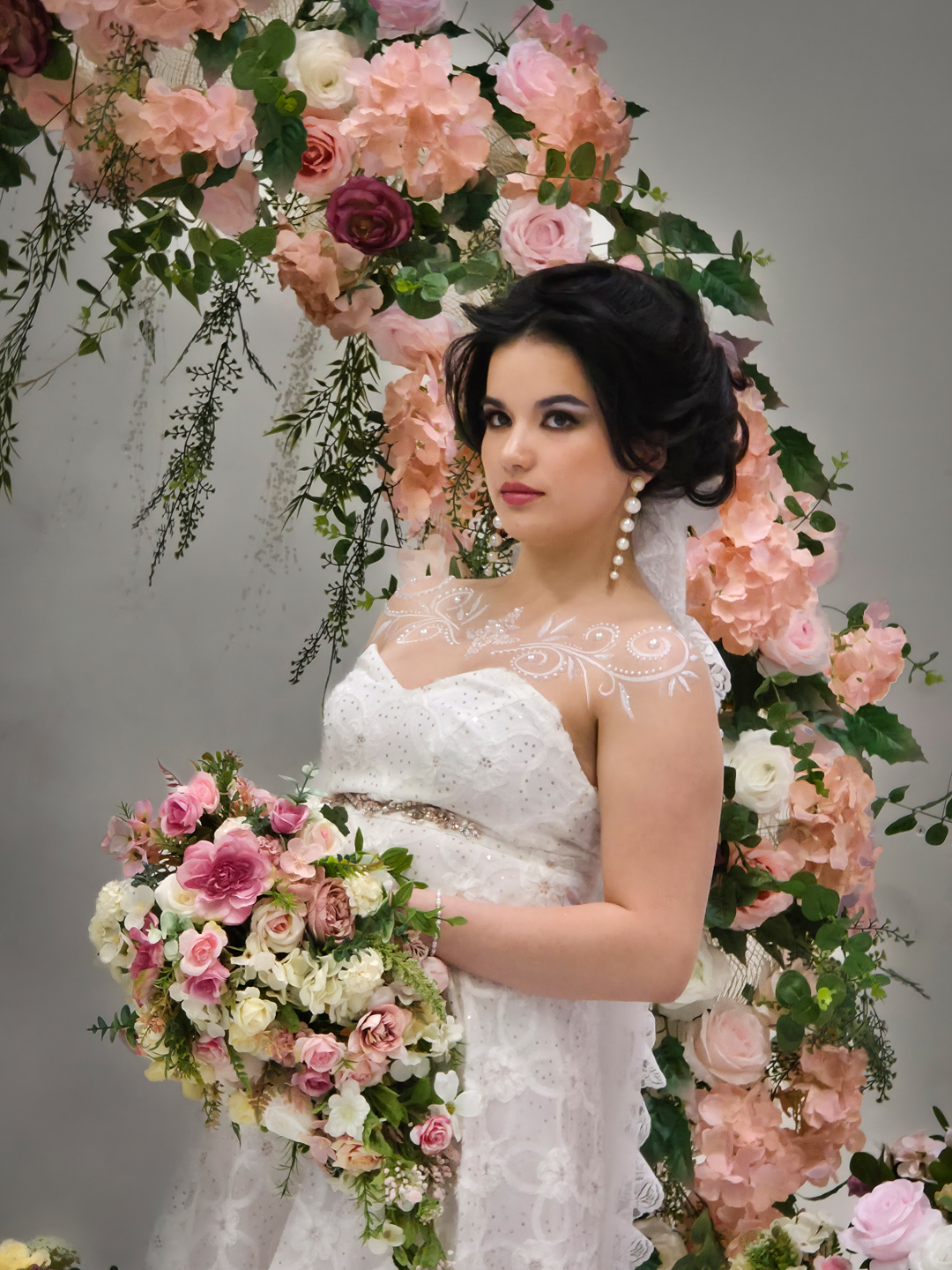 Bride in a white lace dress holding a bouquet in front of a flower arch